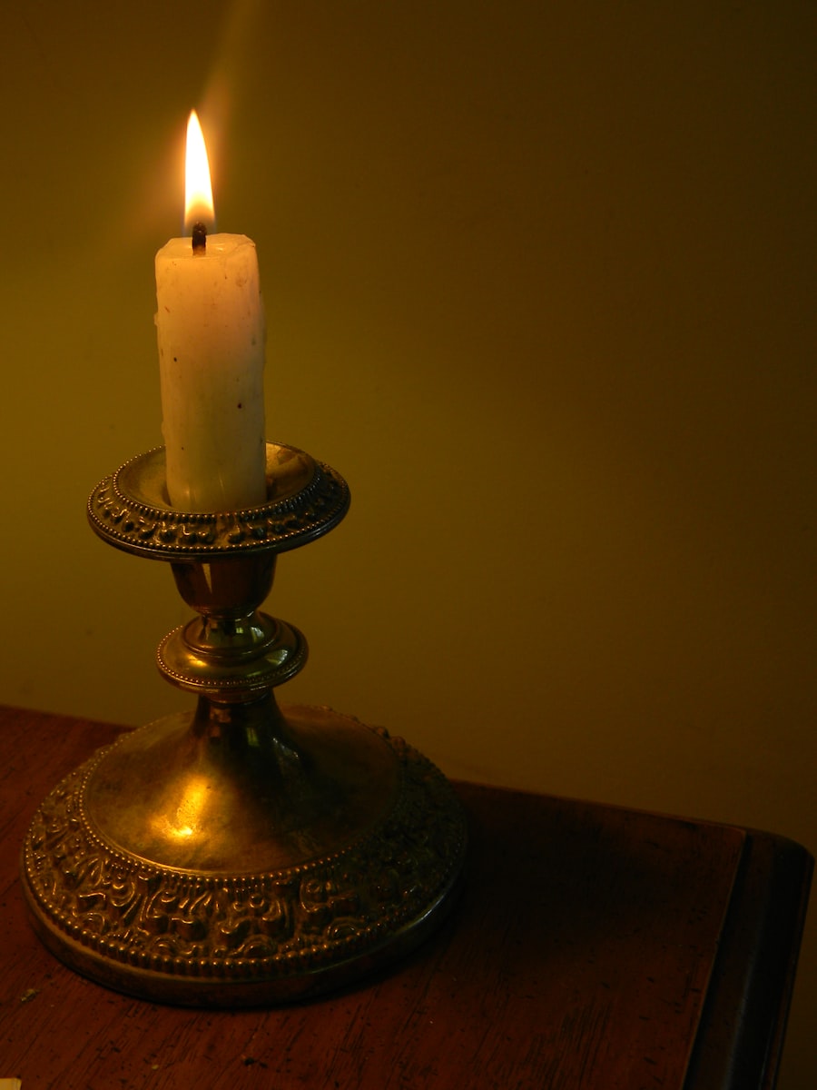 A white candle lit in an ornate brass chamberstick on a wooden table, glowing warm against a deep olive wall.