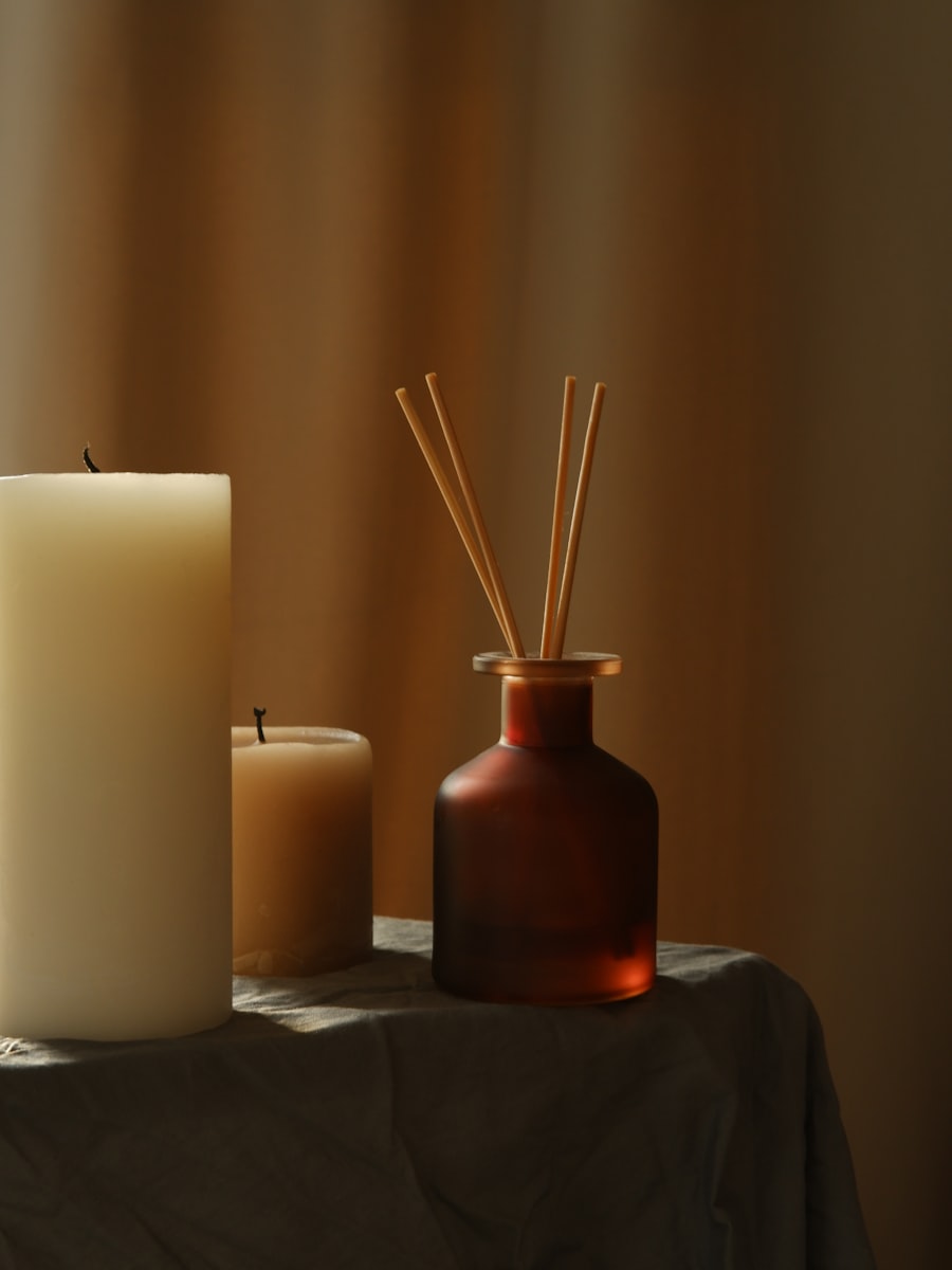 An amber glass reed diffuser with rattan sticks beside two ivory pillar candles in low golden light.