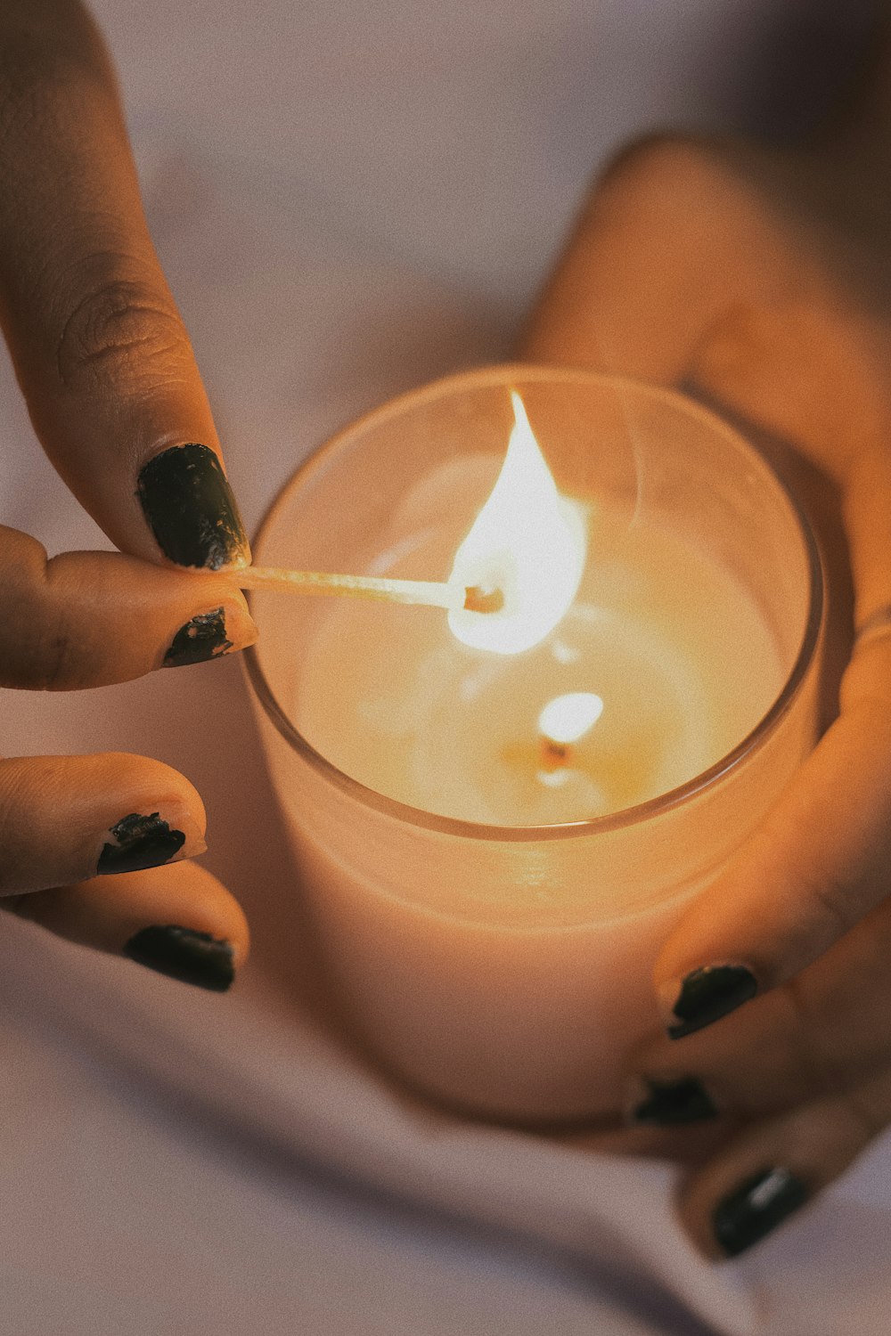Two hands cupping a glass candle and lighting its wick with a wooden match.