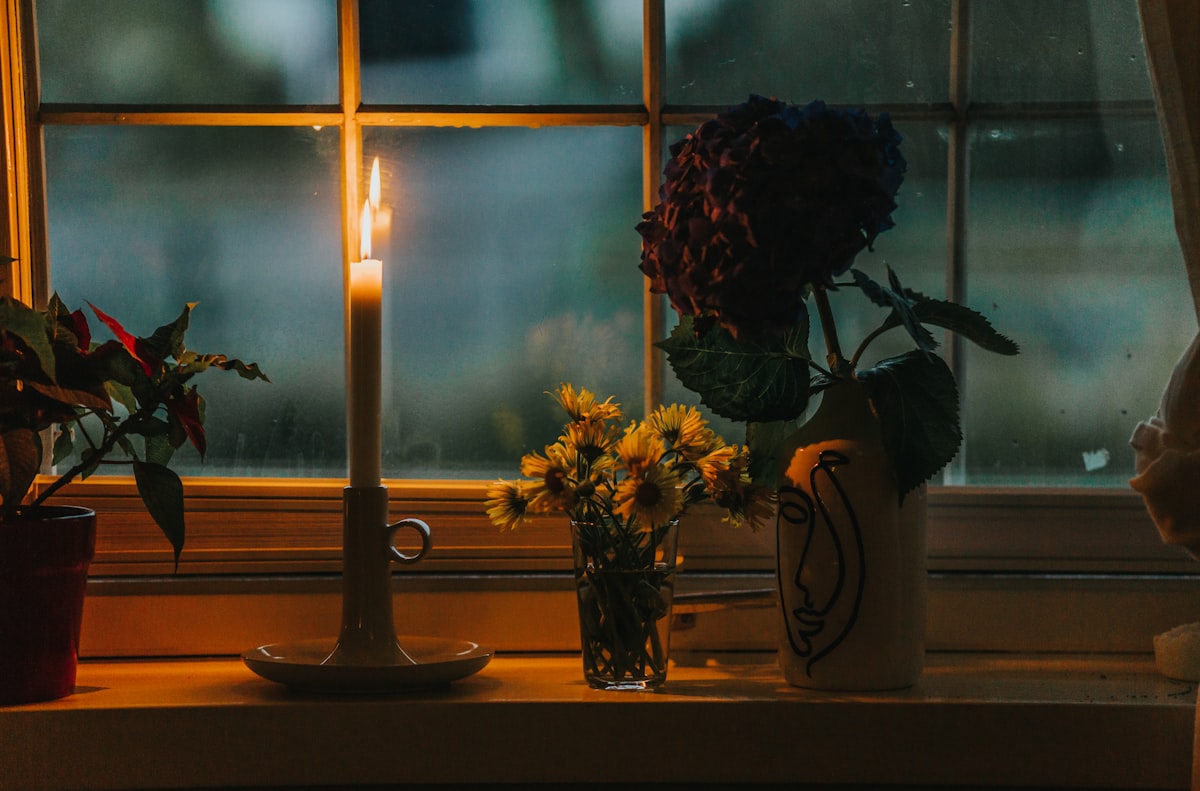 A lit taper candle in a ceramic holder on a windowsill at dusk, beside cut flowers and a rain-flecked window.