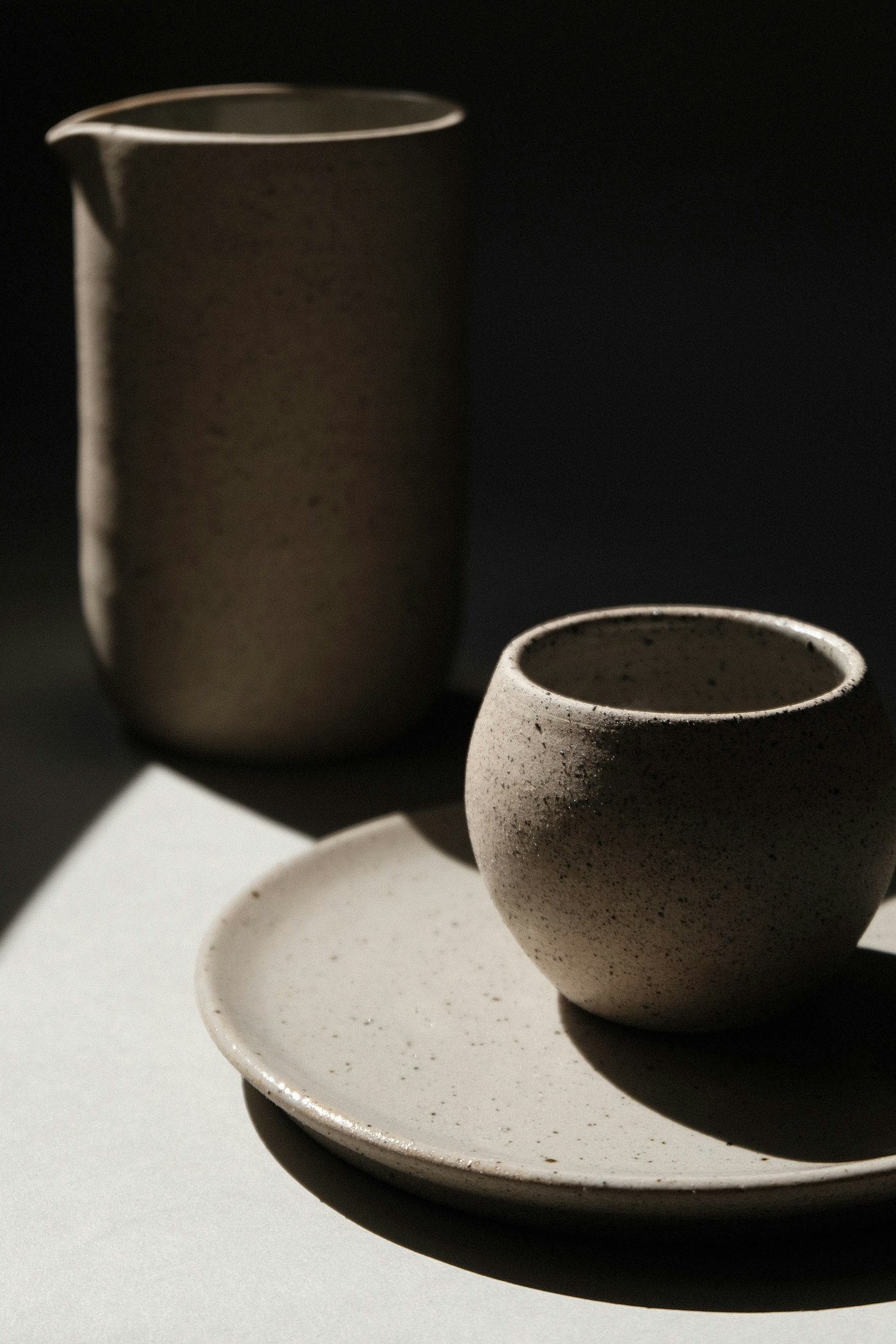 A round speckled stoneware cup on a matching saucer, beside a tall pitcher, in raking light.