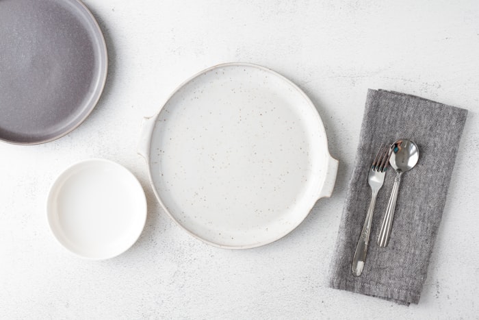 A speckled white stoneware plate and a small bowl laid out beside a grey linen napkin and cutlery.