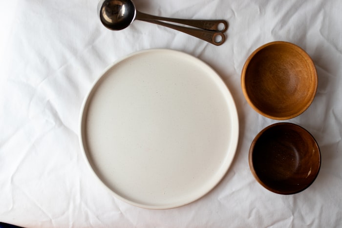 A wide white stoneware plate on linen with two small turned wooden bowls and steel measuring spoons.
