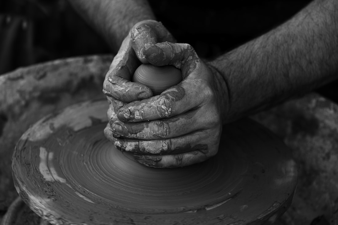 A potter's clay-covered hands closing over a small form spinning on the wheel, photographed in black and white.