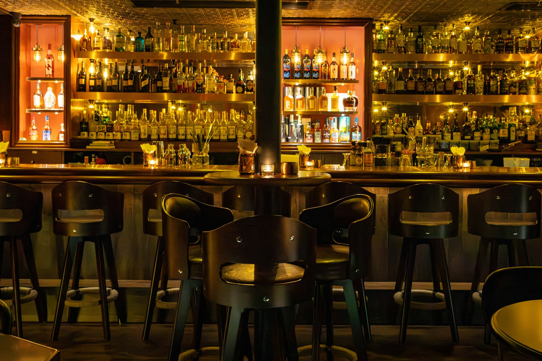 The dimly lit bar of The Alder Room at night — a pressed-tin ceiling, backlit bottle shelves and empty walnut stools along a brass rail.