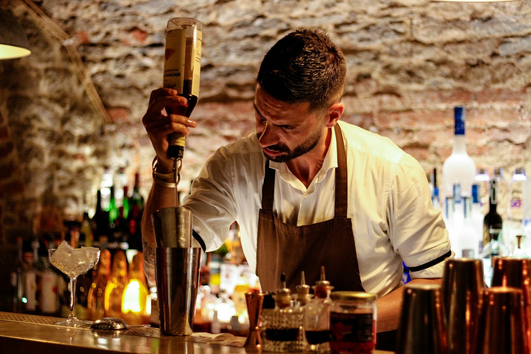 A bartender in a white shirt and brown apron pouring from a bottle into a mixing tin at a stone-walled bar.
