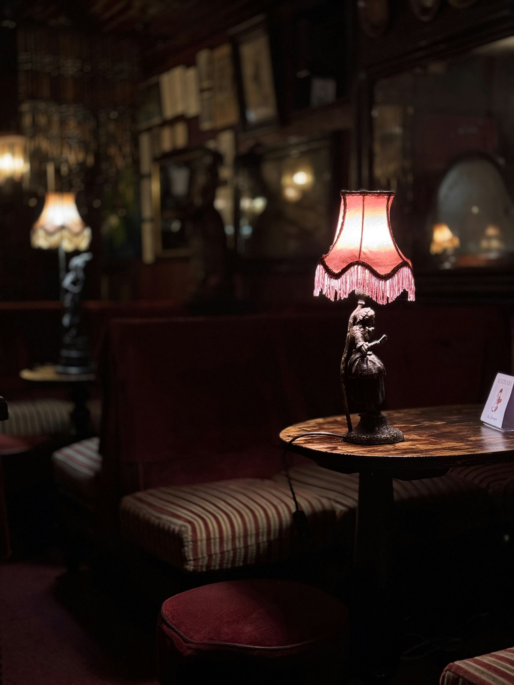 A fringed table lamp glowing on a small round table in a dark bar, with striped banquette seating and framed pictures behind.