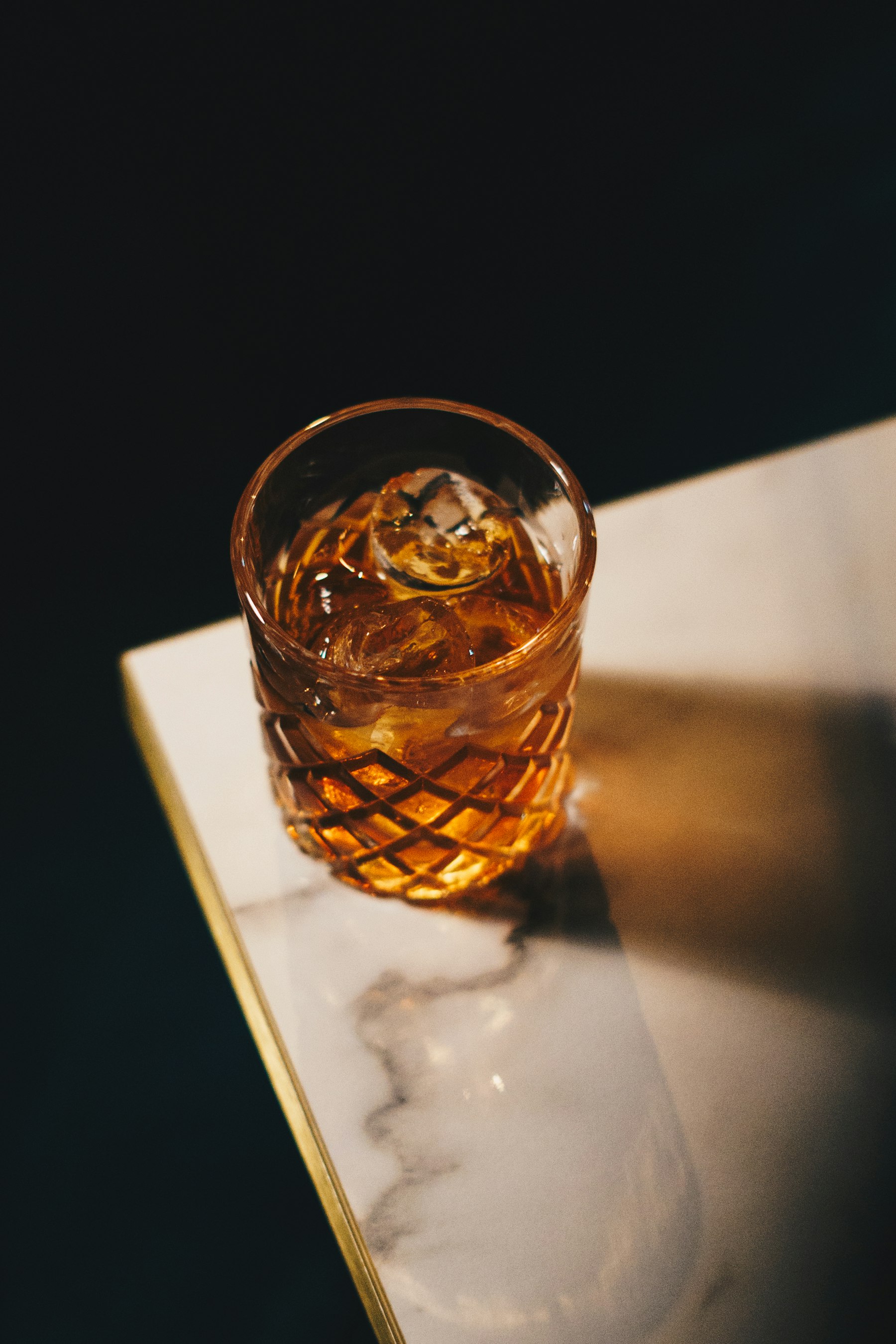 A cut-glass tumbler of amber cocktail with ice, lit by a single warm light on the edge of a marble bar top.