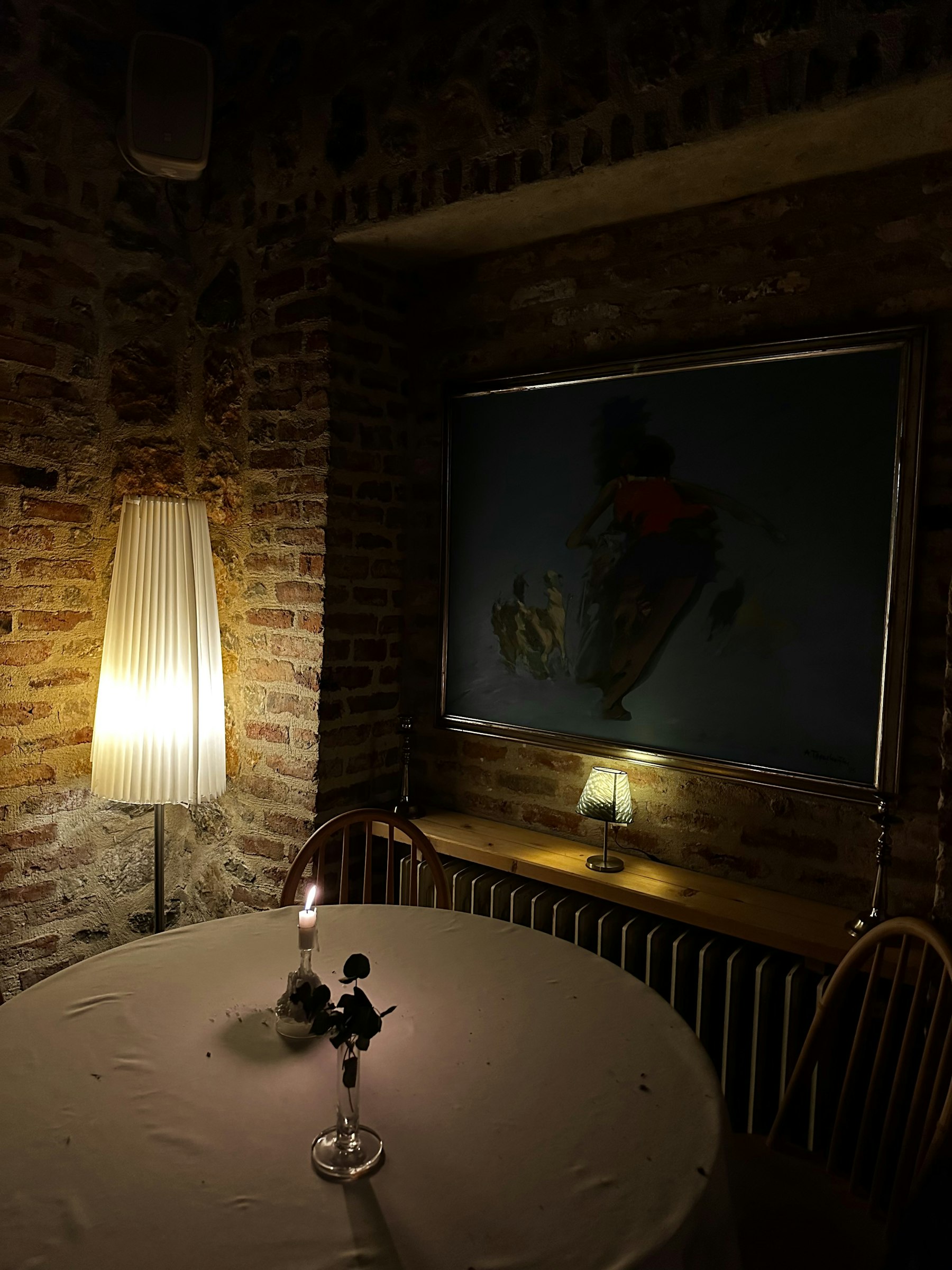 A candlelit round table for two against an exposed brick wall, with a framed painting and a small lamp on the ledge behind.