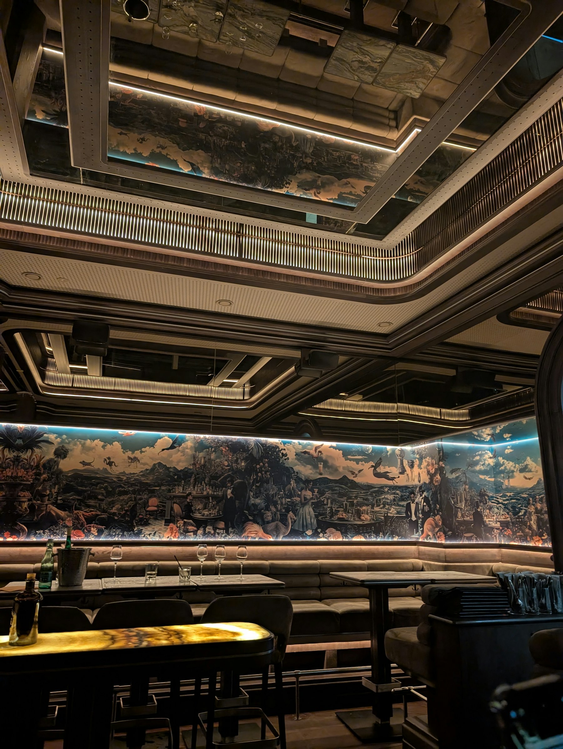 A low-lit lounge with long banquette seating beneath a wide illuminated painted mural and a stepped, mirrored ceiling.