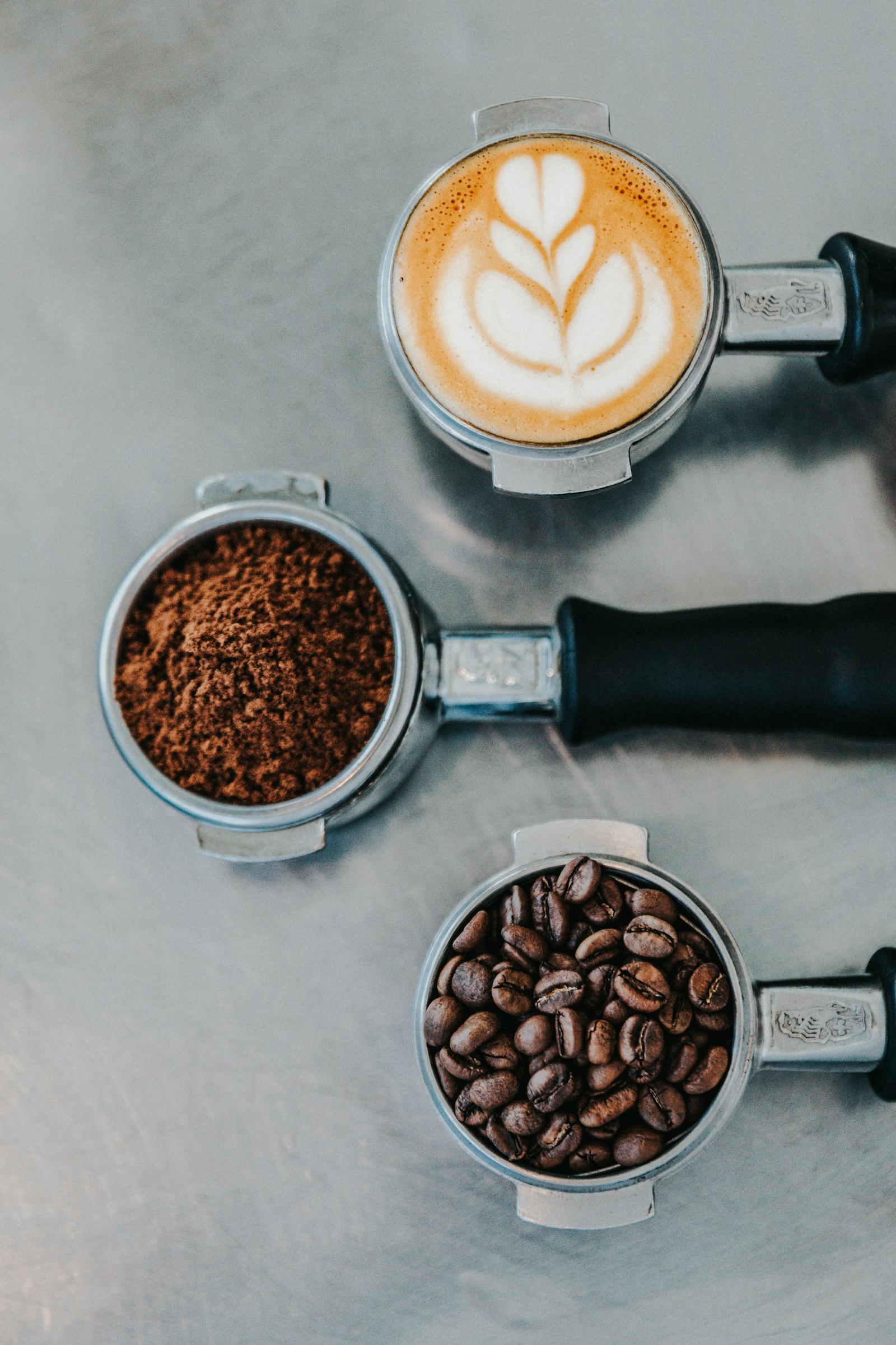 Three espresso portafilters holding whole beans, ground coffee and a finished latte