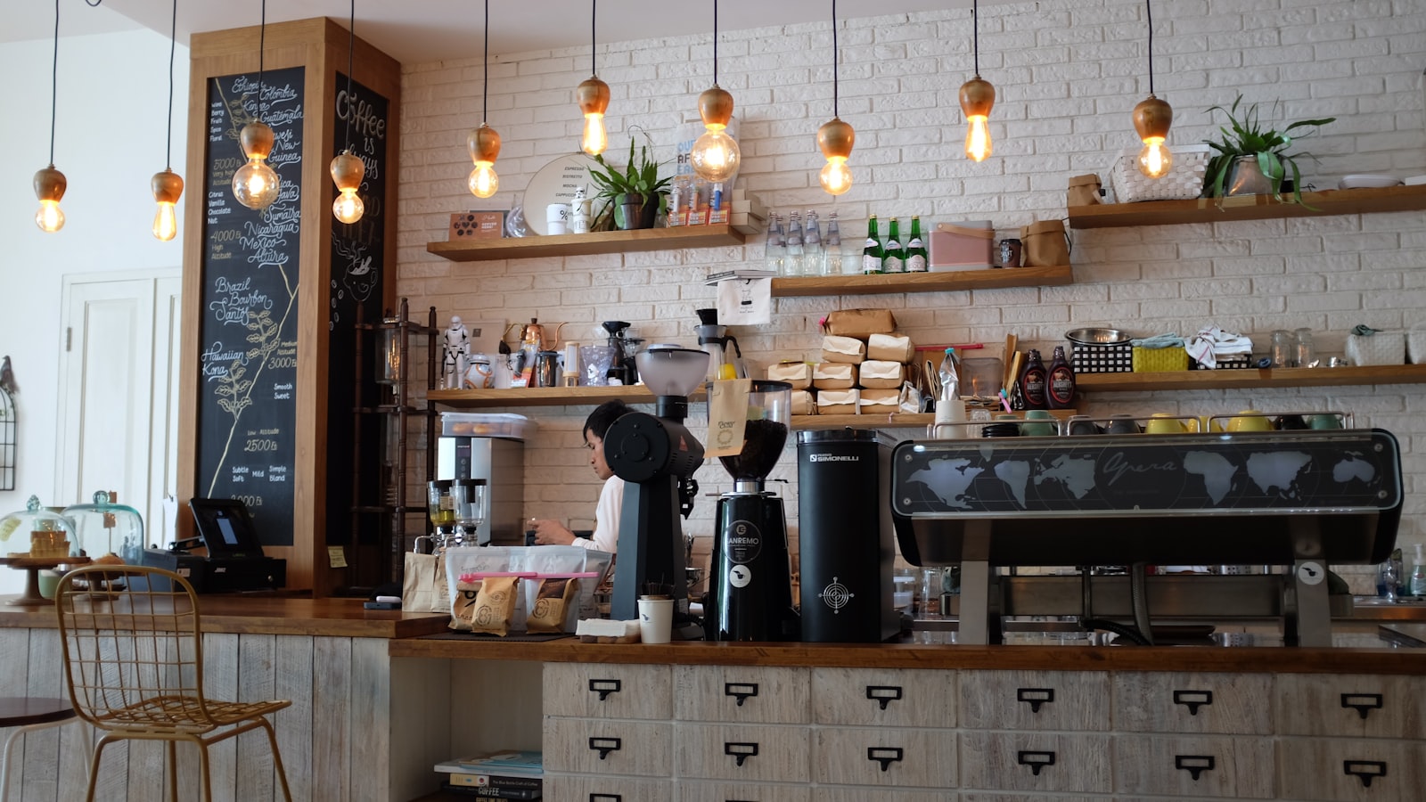 Barista working behind a café bar with an espresso machine, grinders and a chalkboard origin list