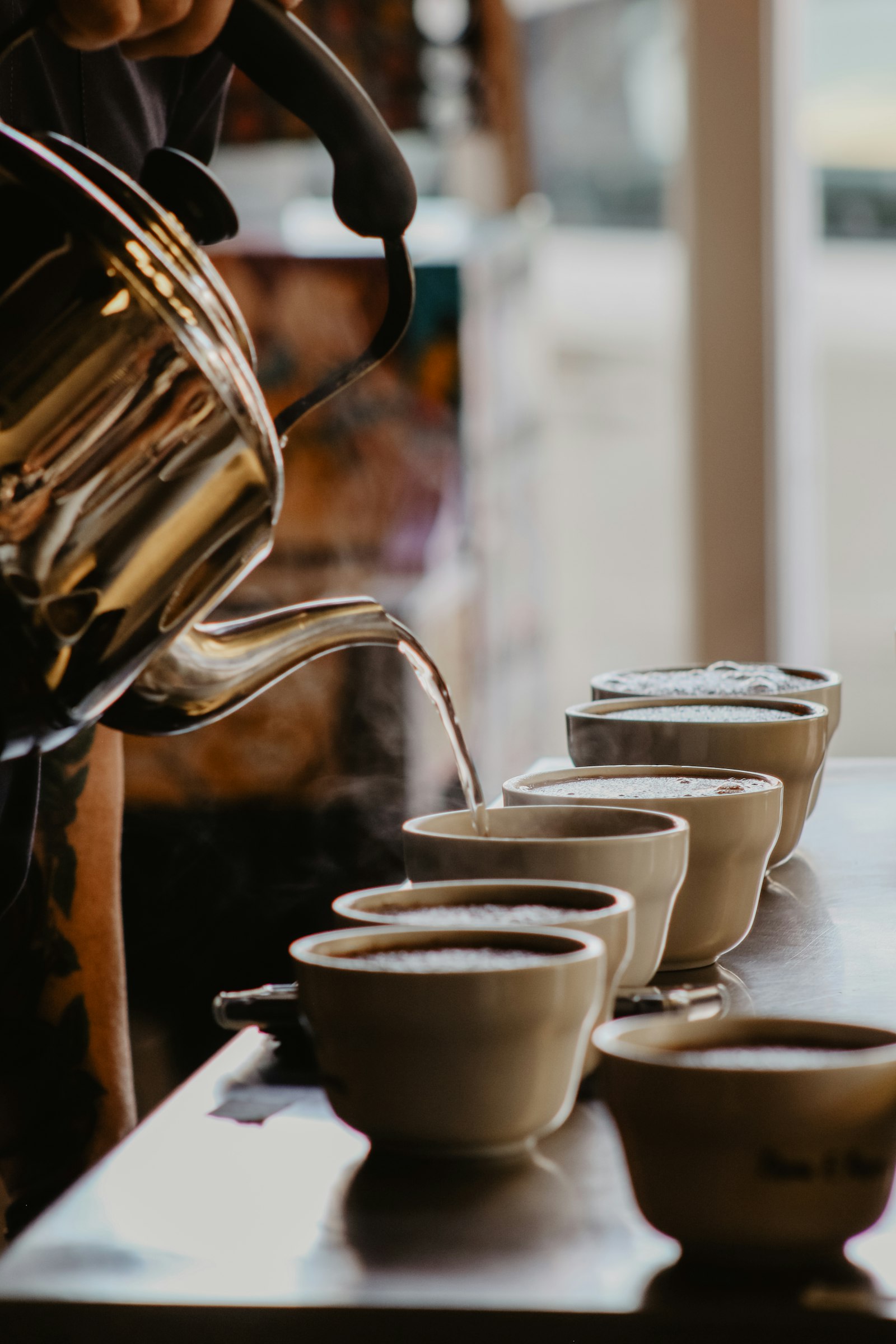 A gooseneck kettle pouring hot water into a row of cupping bowls