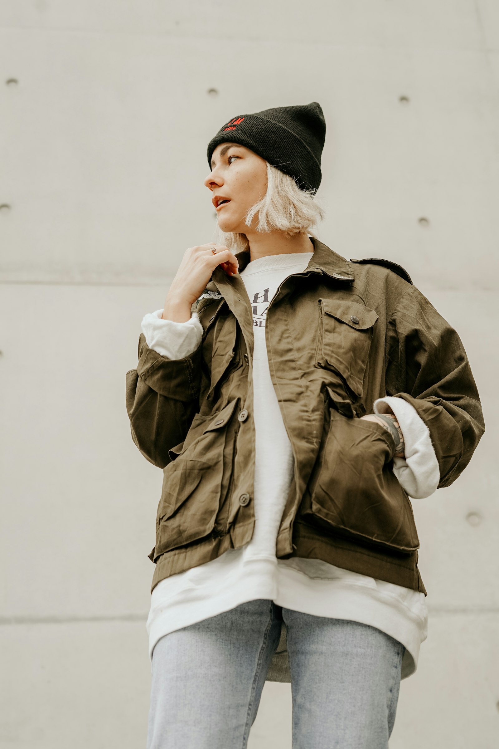 Model in an olive utility jacket layered over an oversized tee, wearing a black beanie against concrete