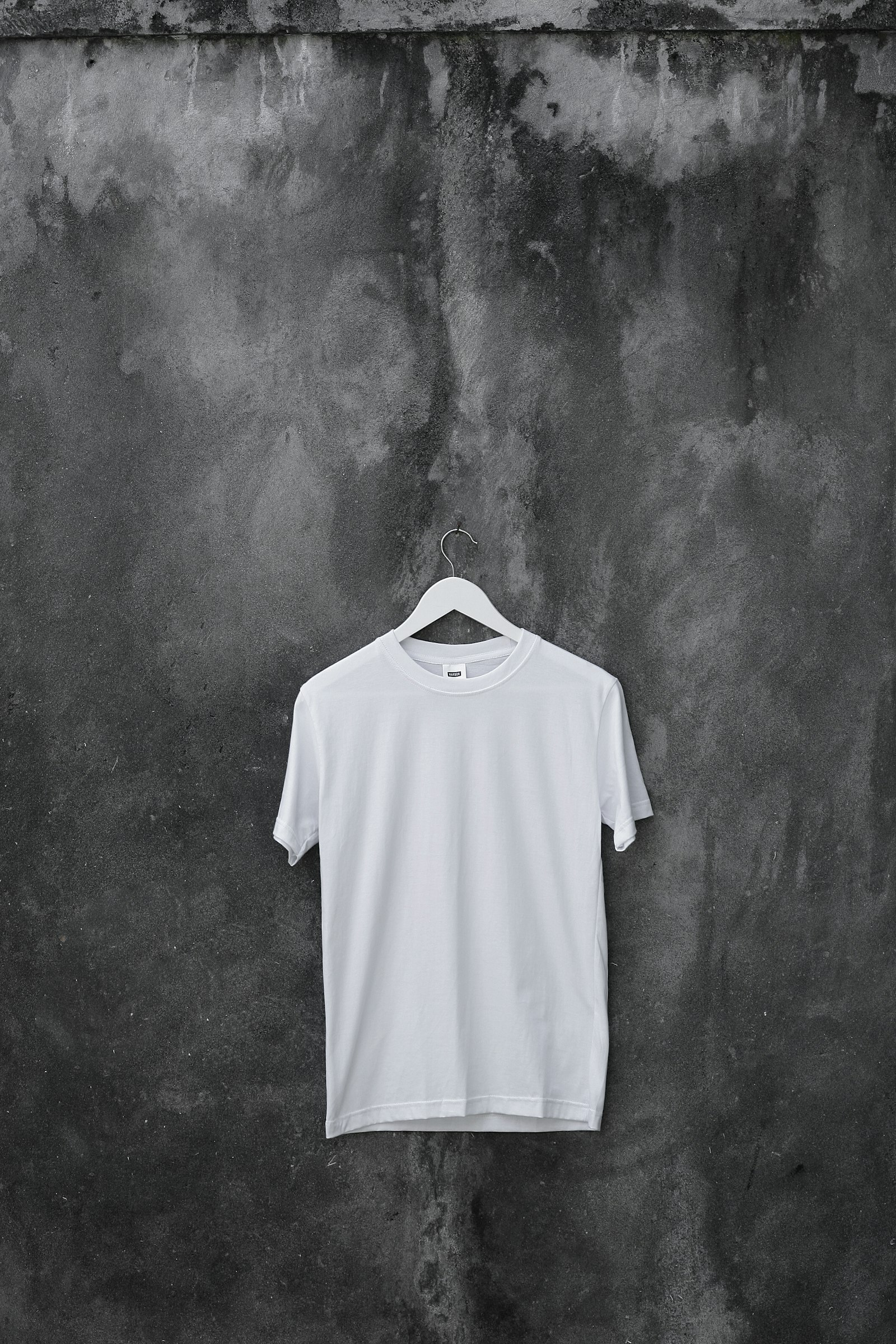 Heavyweight blank tee hanging on a slim hanger against a raw concrete wall
