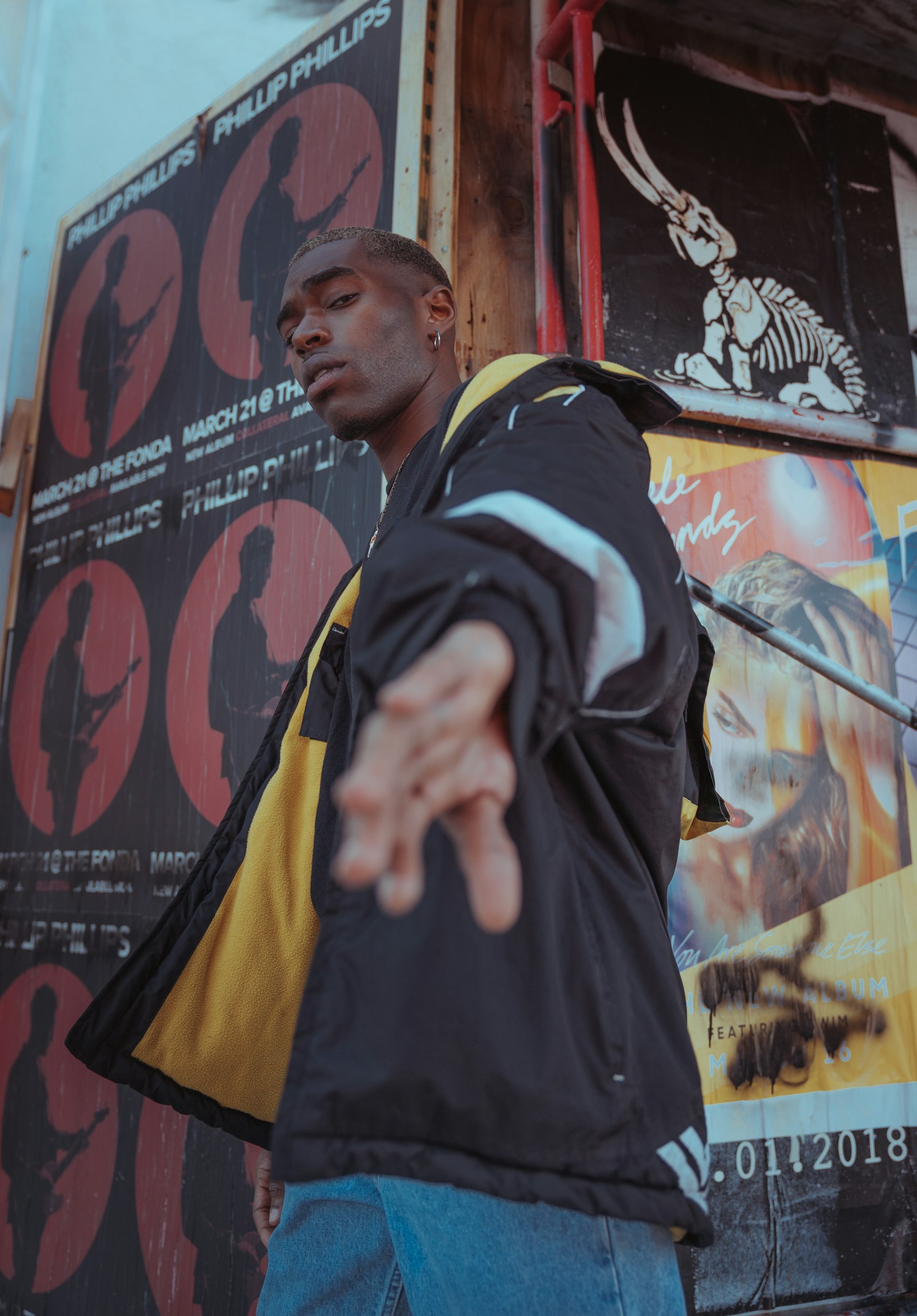 Model in a black and yellow panelled shell jacket standing against a wall of layered gig posters