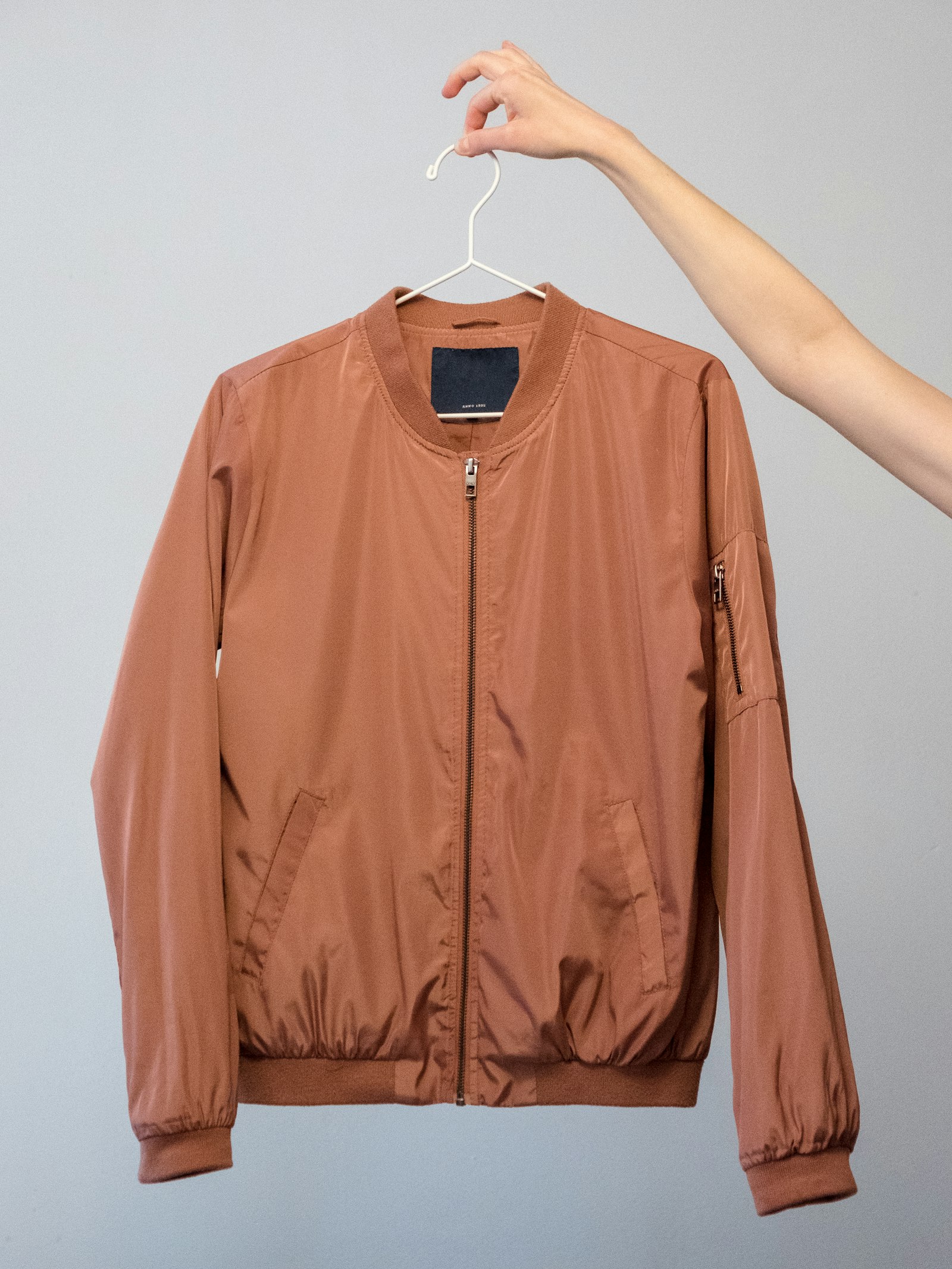 Rust-coloured nylon bomber jacket held up on a white hanger against a grey wall