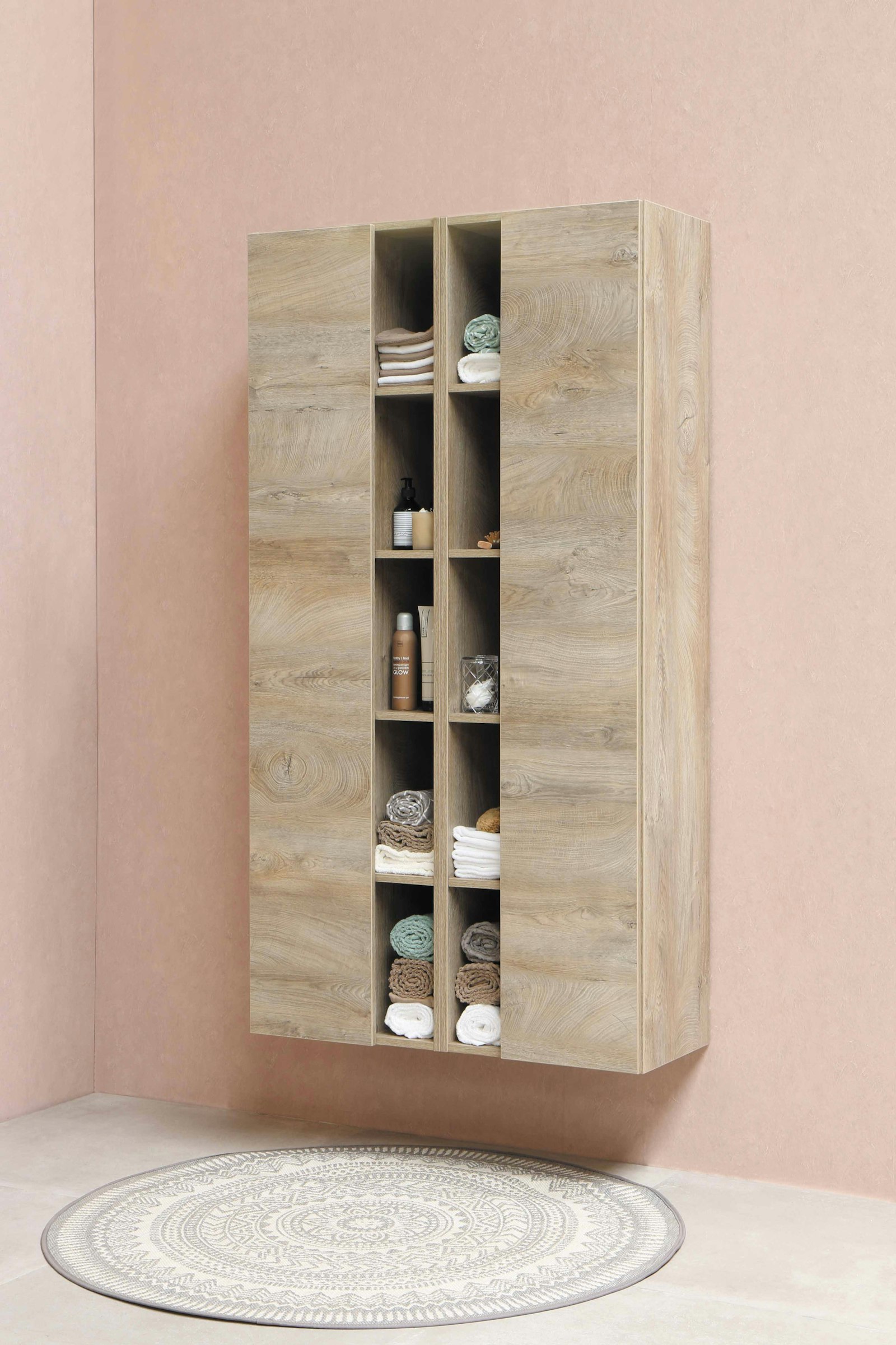 Tall oak shelving unit with open cubbies holding folded linens, mounted on a clay-pink wall