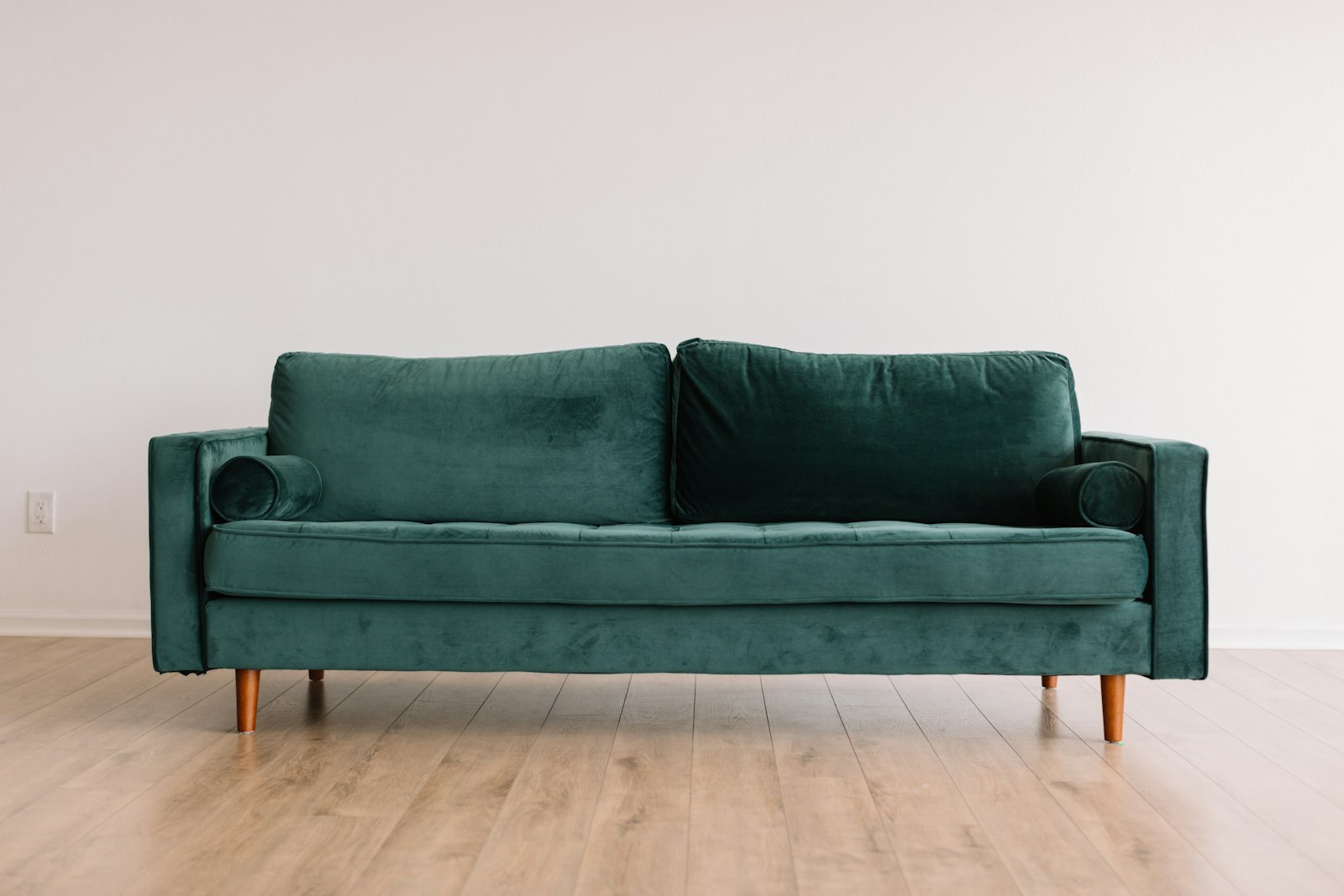 Deep green velvet three-seater sofa with bolster cushions and turned wooden feet on a pale wood floor