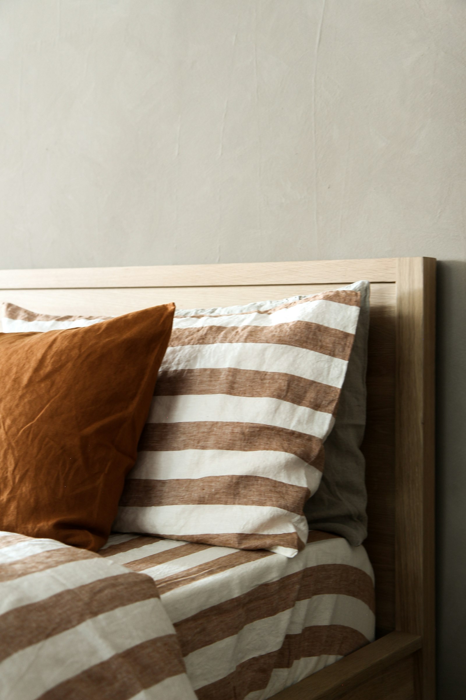 Pale oak bed headboard dressed in rust-striped washed linen bedding and a burnt-orange linen pillow