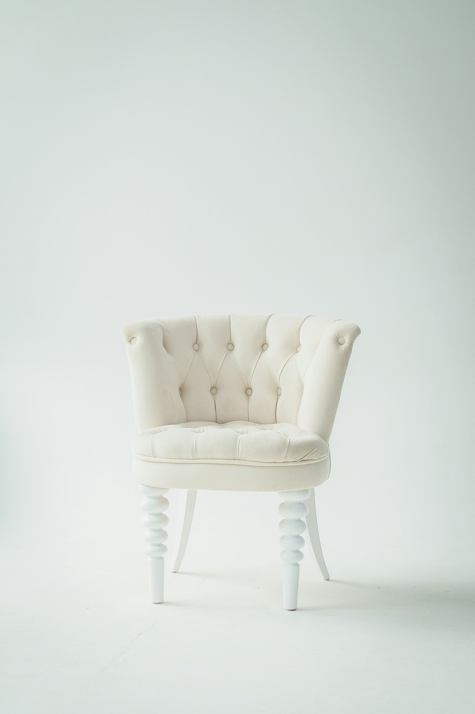 Cream tufted occasional chair with turned white legs on a plain studio backdrop