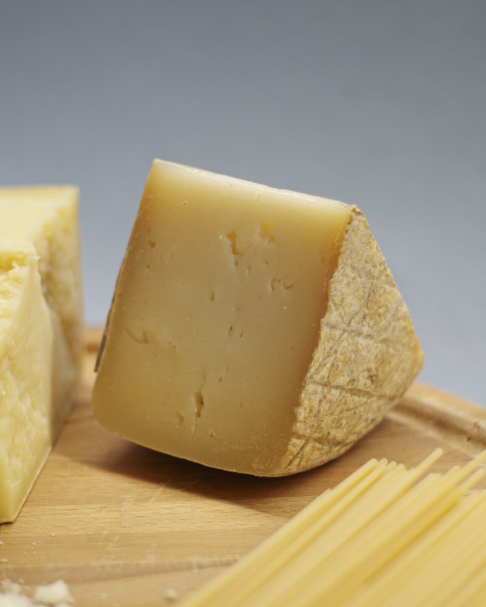 A wedge of hard farmhouse cheese on a wooden board beside dried pasta