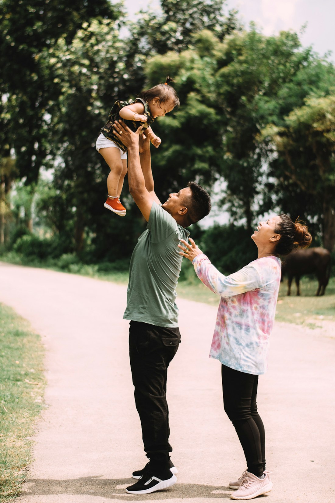 A father lifting his toddler in the air on a park path while her mother laughs beside them
