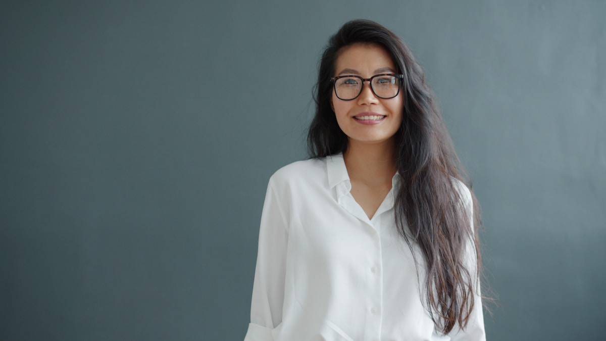 Dr Mei-Lin Reyes, medical director — studio portrait of a woman in glasses and a white shirt on a grey ground.