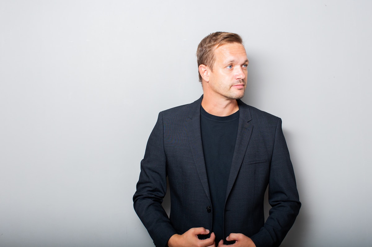 Dr Anders Holm, aesthetic physician — studio portrait of a man in a dark jacket on a pale grey ground.