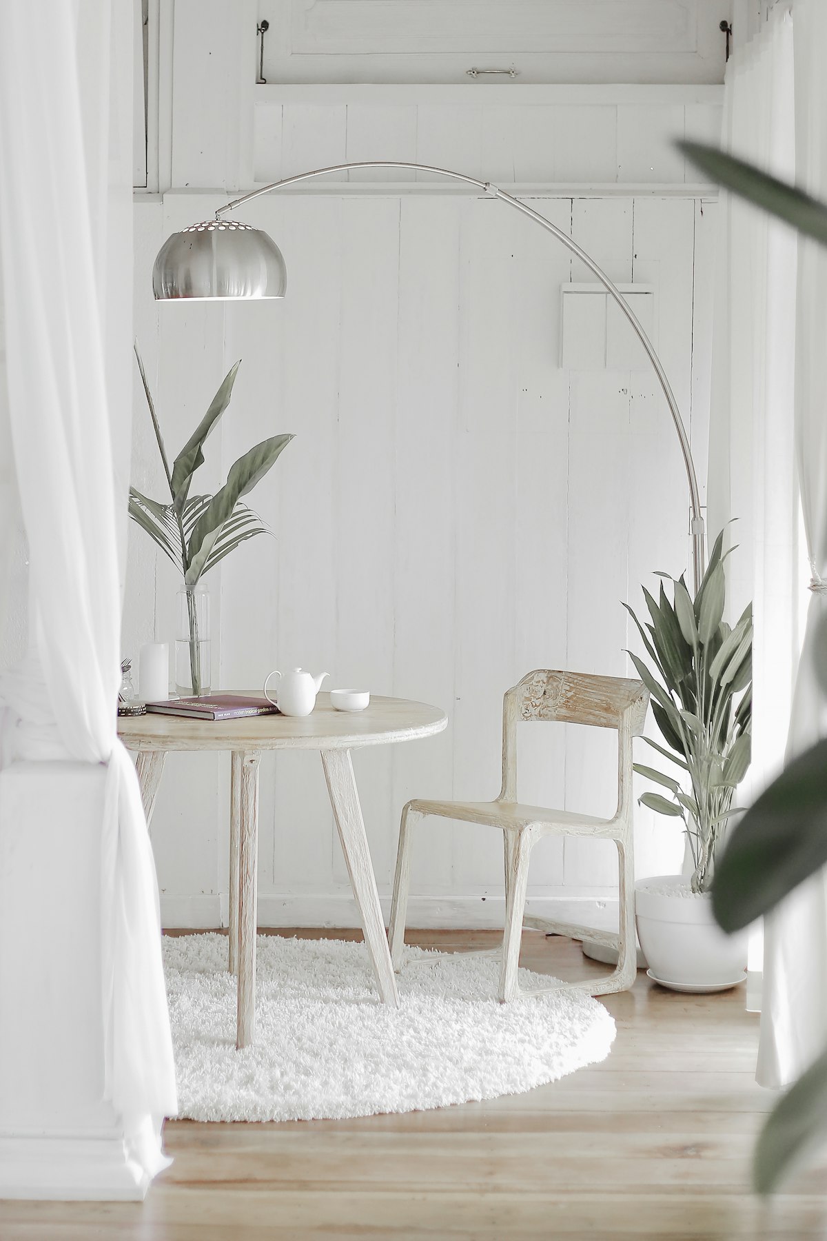 A quiet, sunlit corner of a home with a small round table, a wooden chair, a plant and soft white curtains