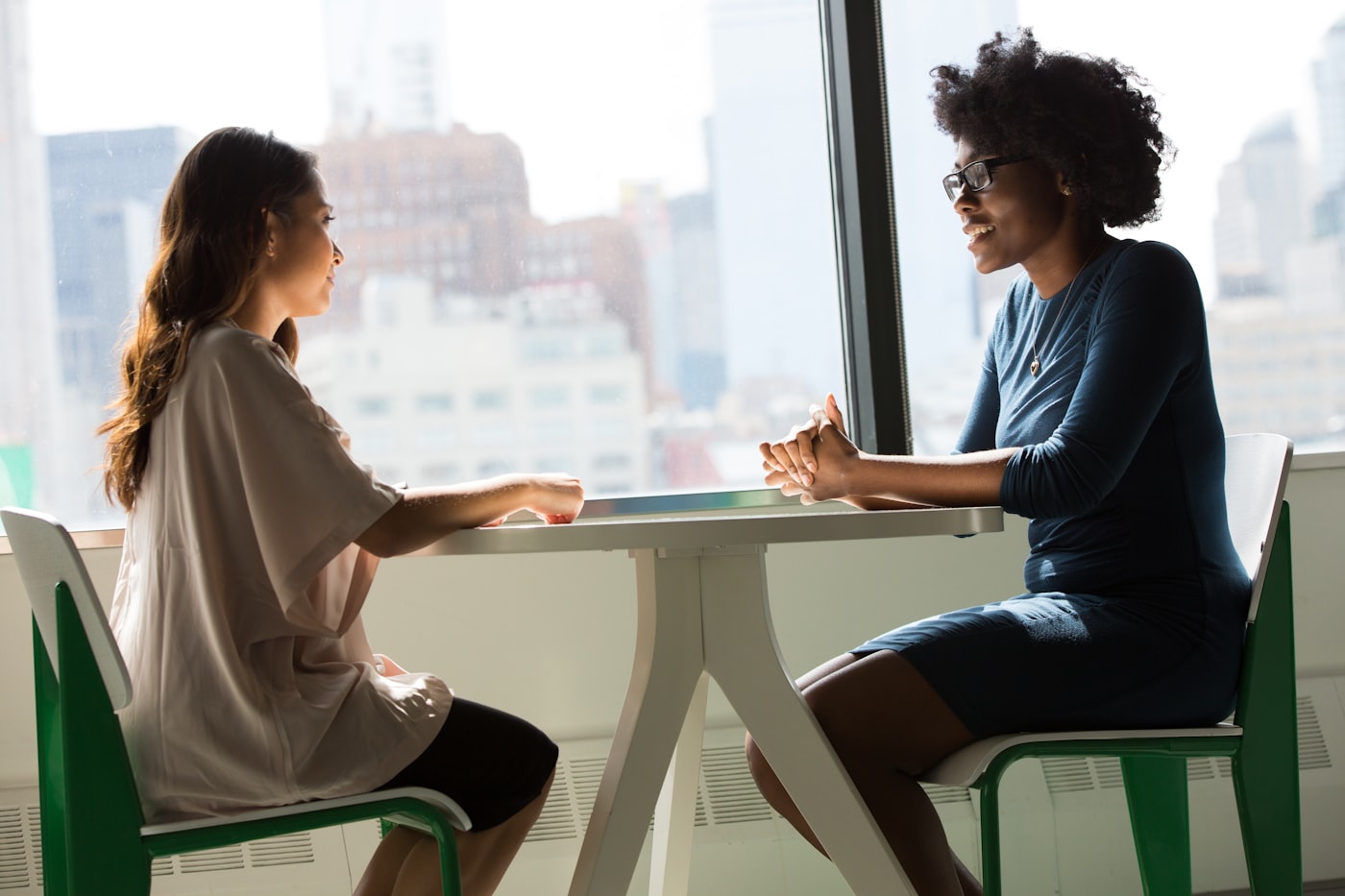Two women sitting across a small table by a bright window, one listening closely as the other speaks