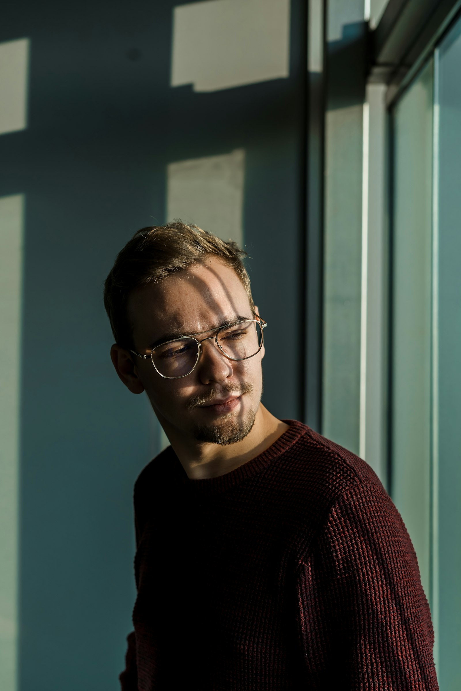 Young man in thin metal-framed spectacles beside a window, hard sunlight falling across his face and a teal wall behind