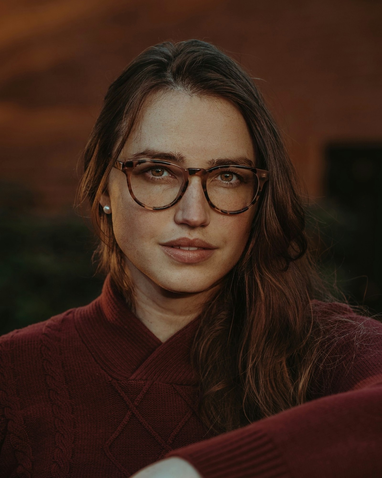 Woman in tortoiseshell round spectacles and a maroon cable-knit jumper, warm blurred background