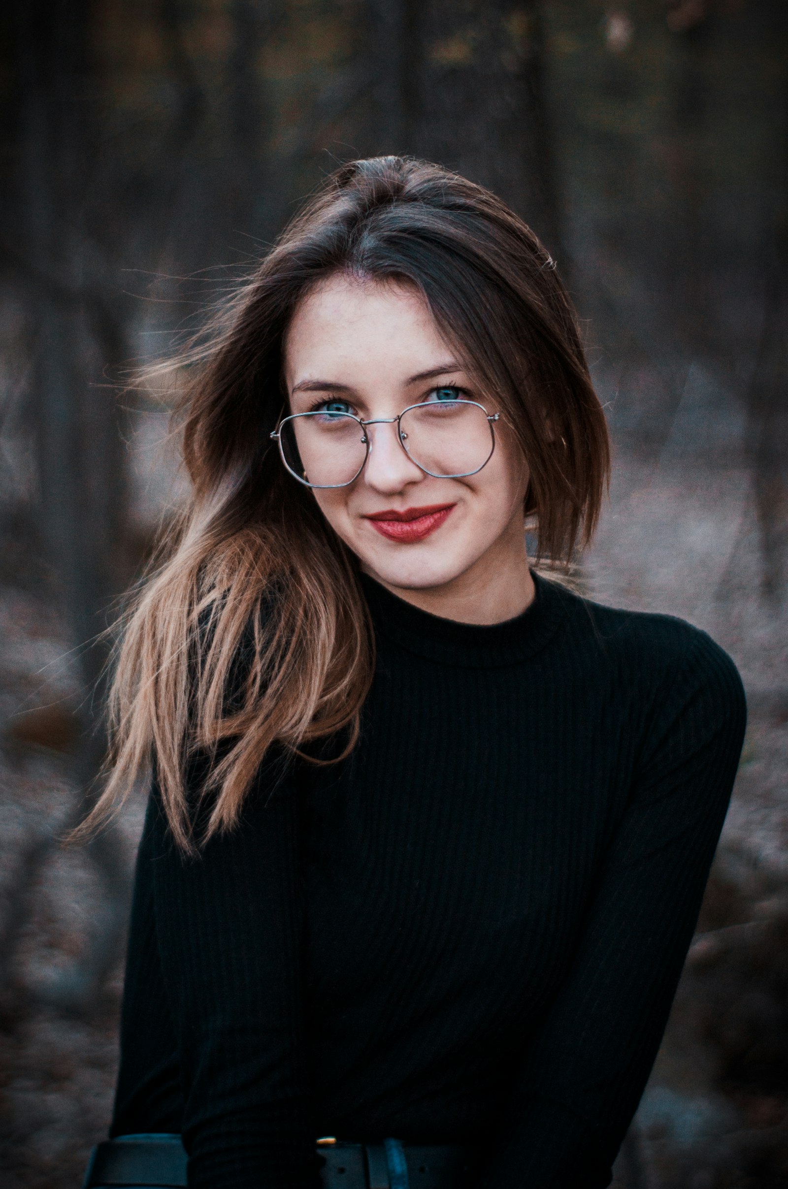 Woman in round wire-framed spectacles and a black roll-neck, bare woodland out of focus behind her