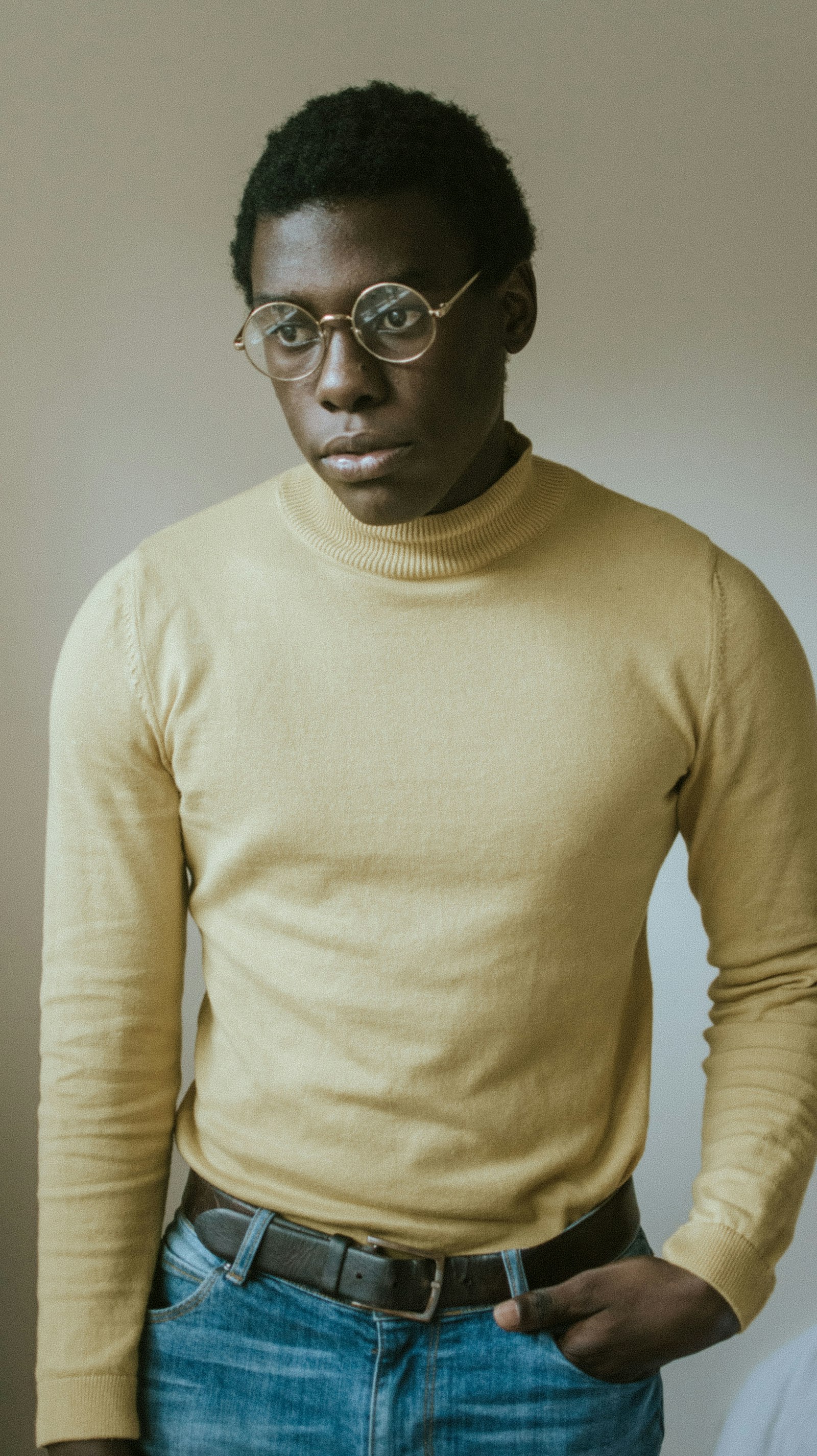 Man in a pale yellow turtleneck wearing round gold wire-framed spectacles, standing against a plain wall