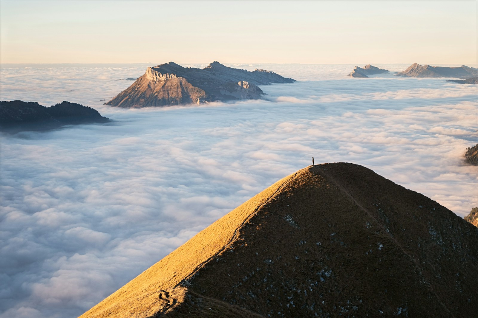 A single figure standing on a narrow sunlit ridge above a vast sea of cloud at sunrise