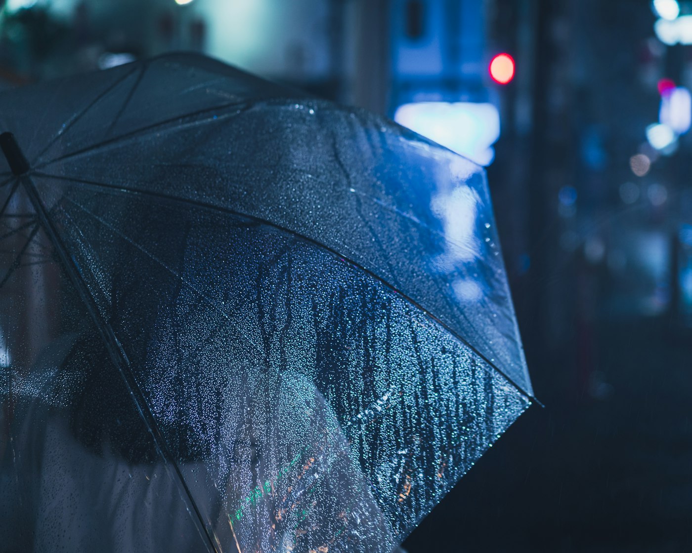Clear umbrella beaded with rain at night, city lights blurred to blue and red bokeh behind it