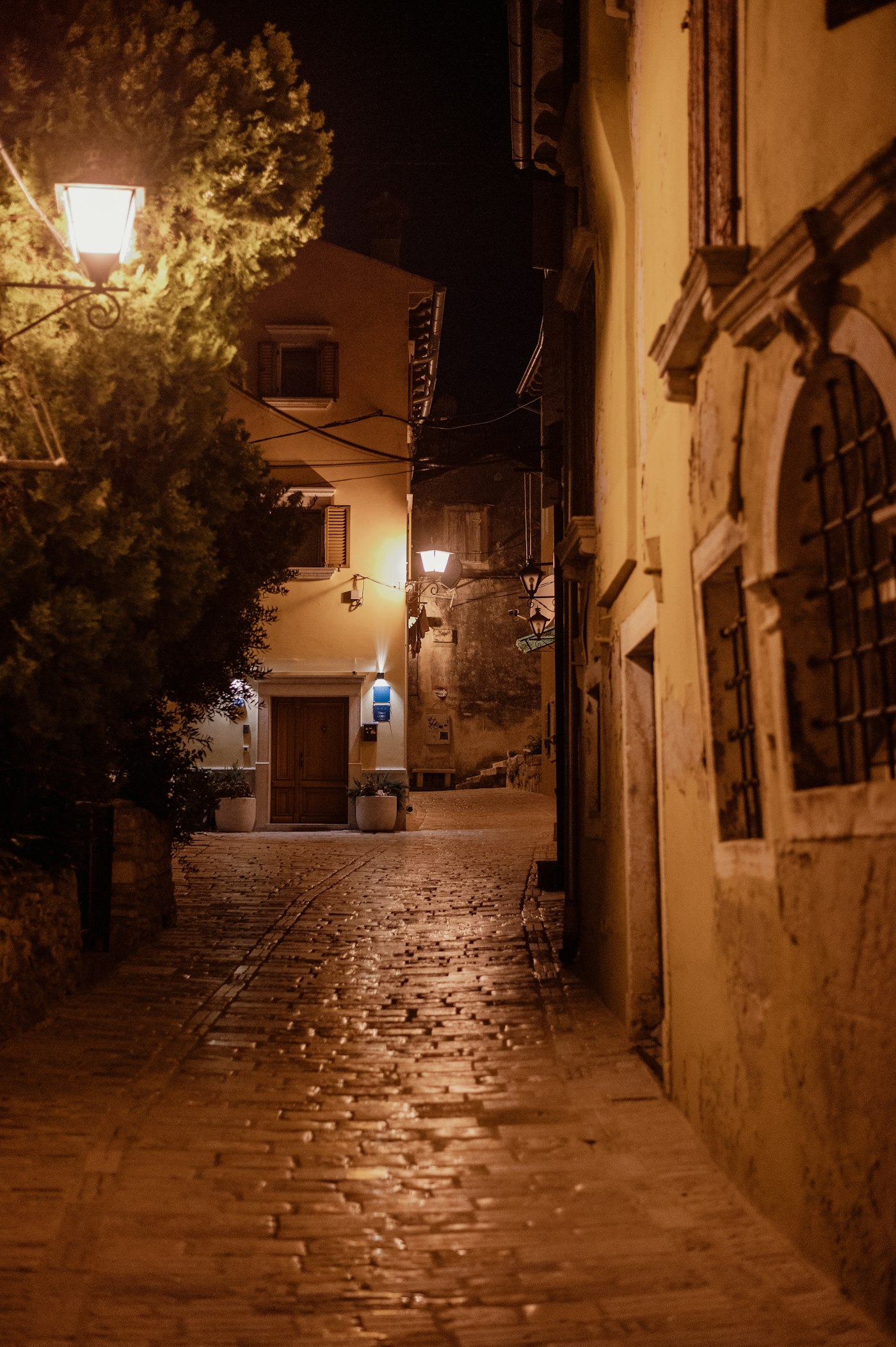 Empty cobbled lane at night, wet stone reflecting a chain of amber lamps