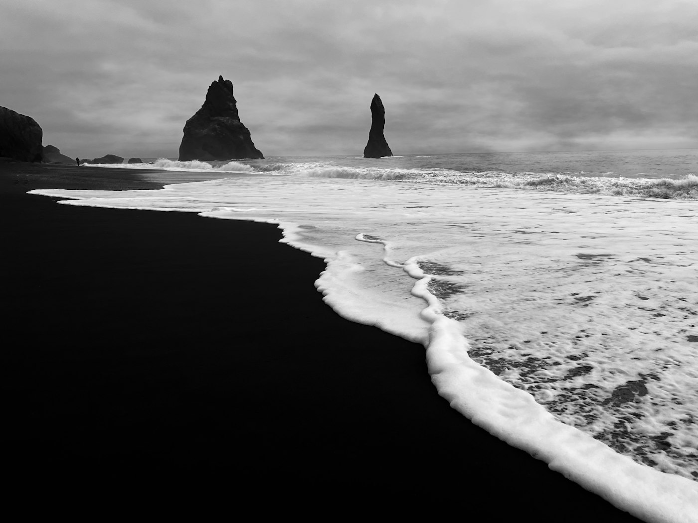 Black-sand shoreline in monochrome, sea stacks standing in surf under a flat sky