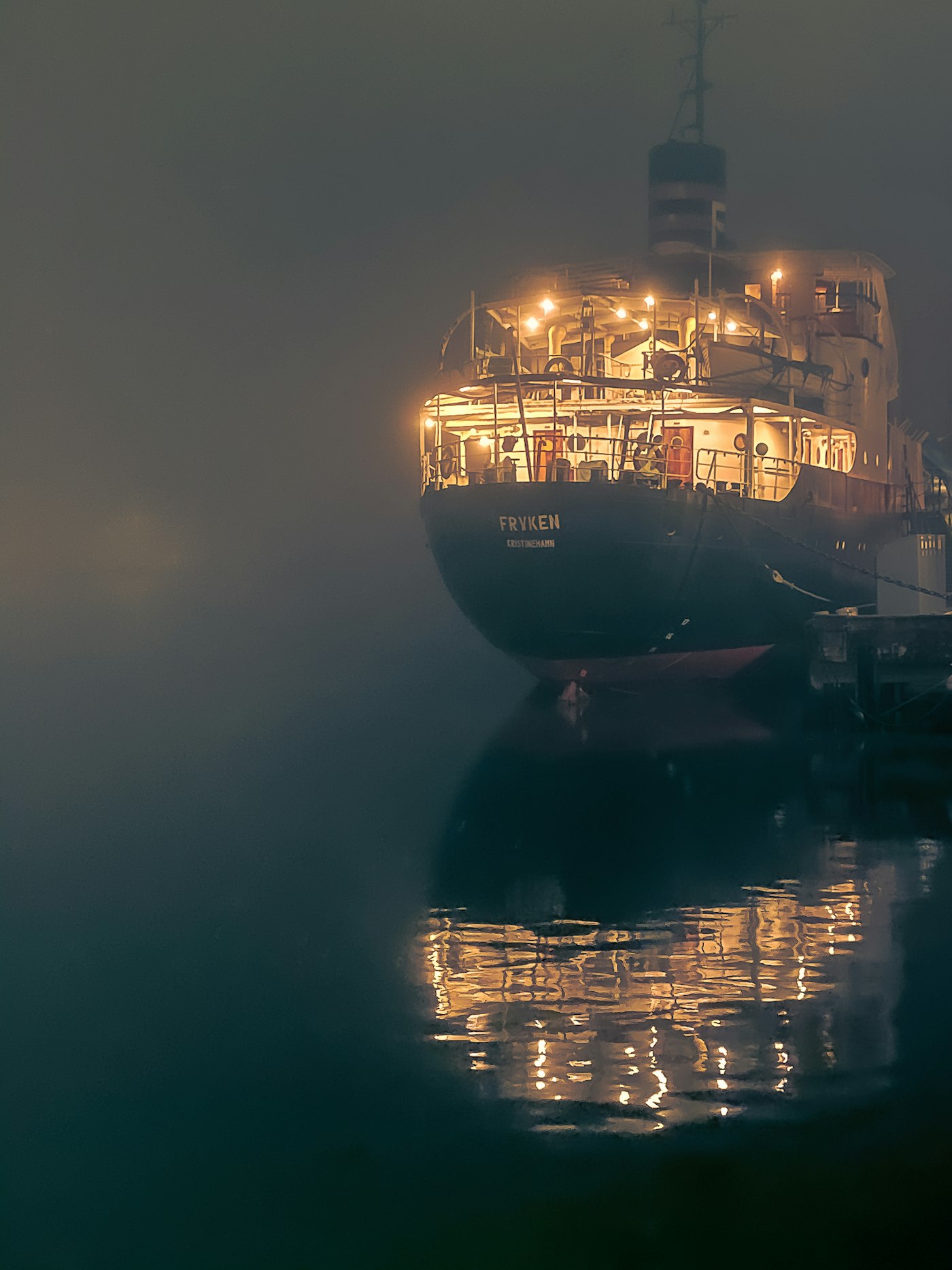 Lit ship moored in harbour fog, its deck lamps reflected in still black water