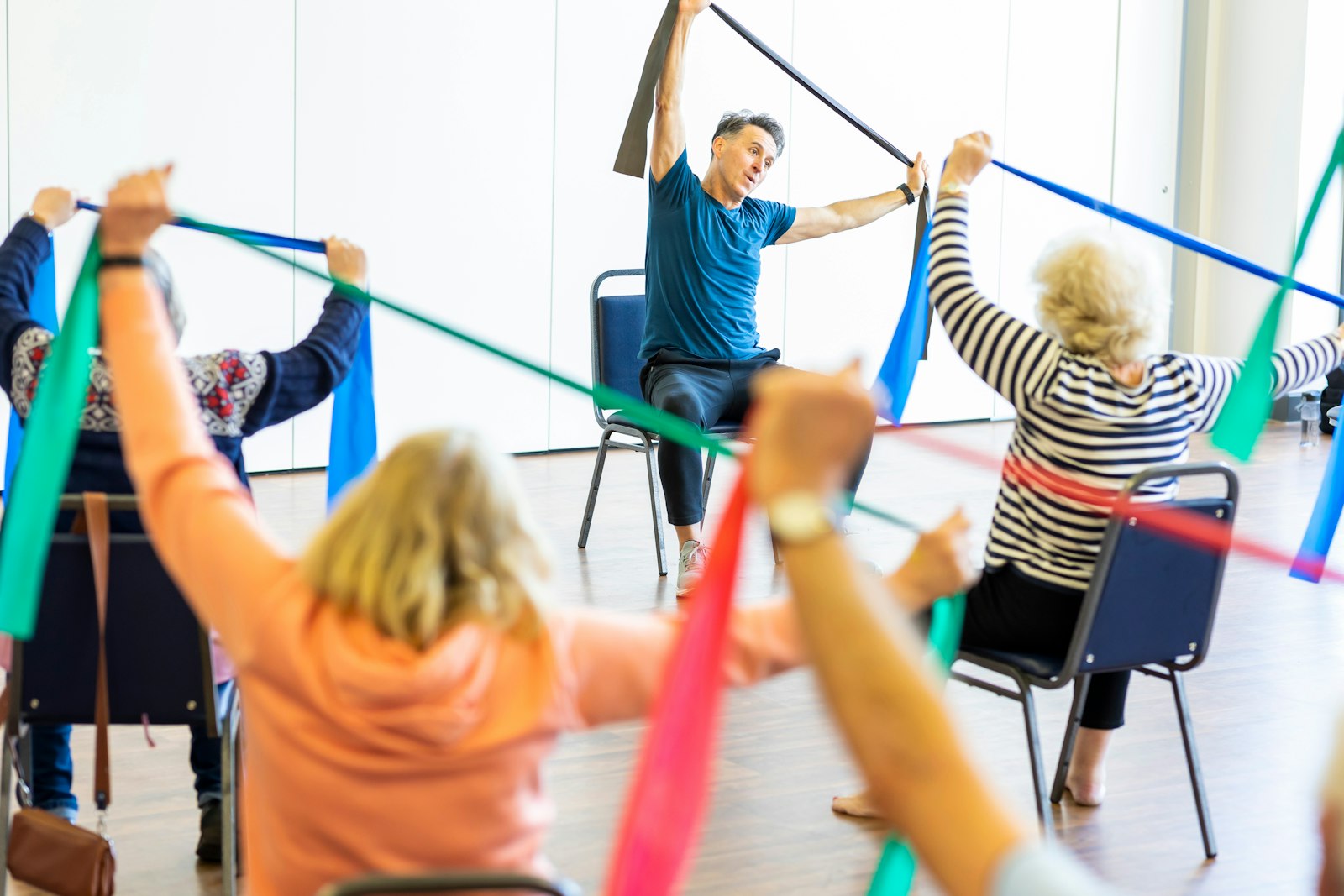 A seated resistance-band class in progress: older adults raising coloured therapy bands overhead while an instructor demonstrates from the front.