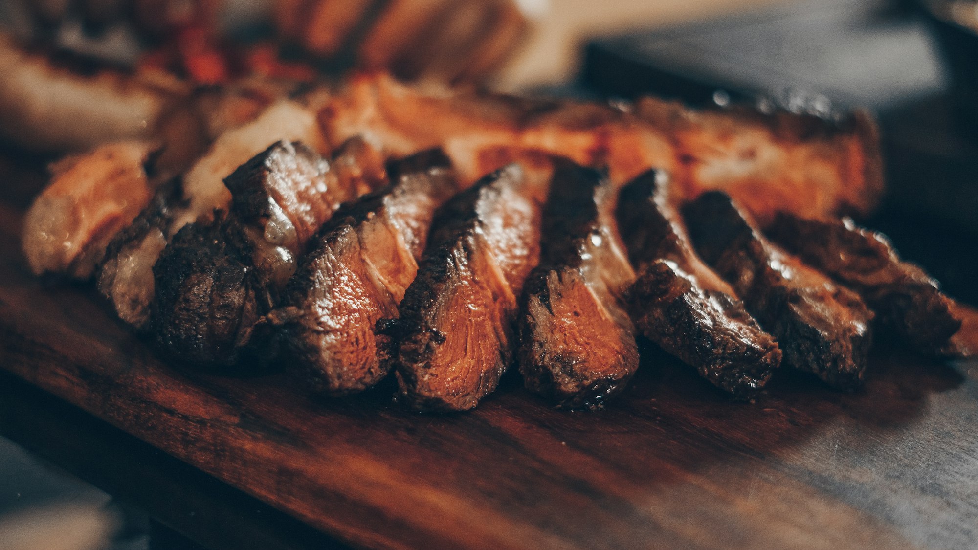 Oak-smoked short rib carved into slices on a charred wooden board, lit by low candlelight