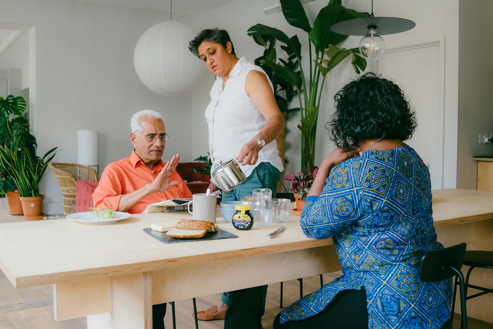An older man seated at a long dining table gestures in conversation while a staff member pours from a teapot and another resident sits opposite.