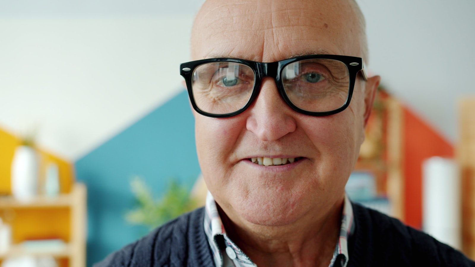 Close portrait of an older man in heavy black-framed glasses and a dark cardigan, smiling at the camera.