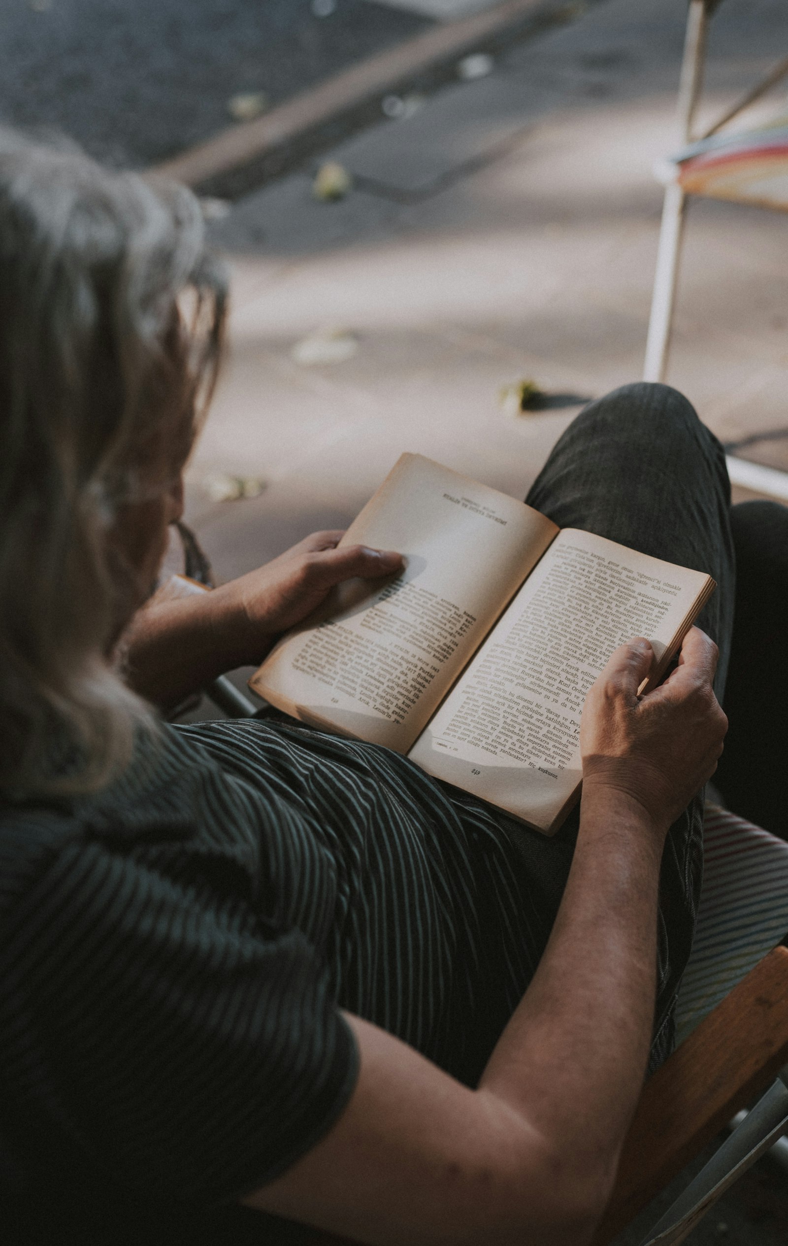 An older woman with grey hair, seen over her shoulder, reading an open hardback book on her lap.