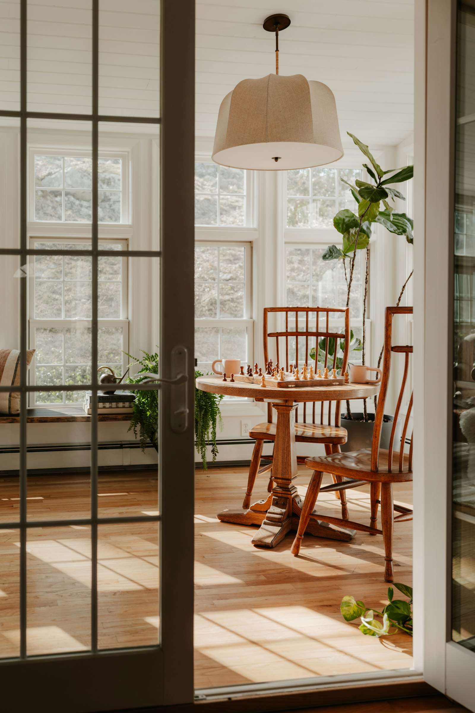 A sunroom seen through open glass doors, with a chess set mid-game on a round wooden table, spindle-back chairs and low afternoon light across the floorboards.