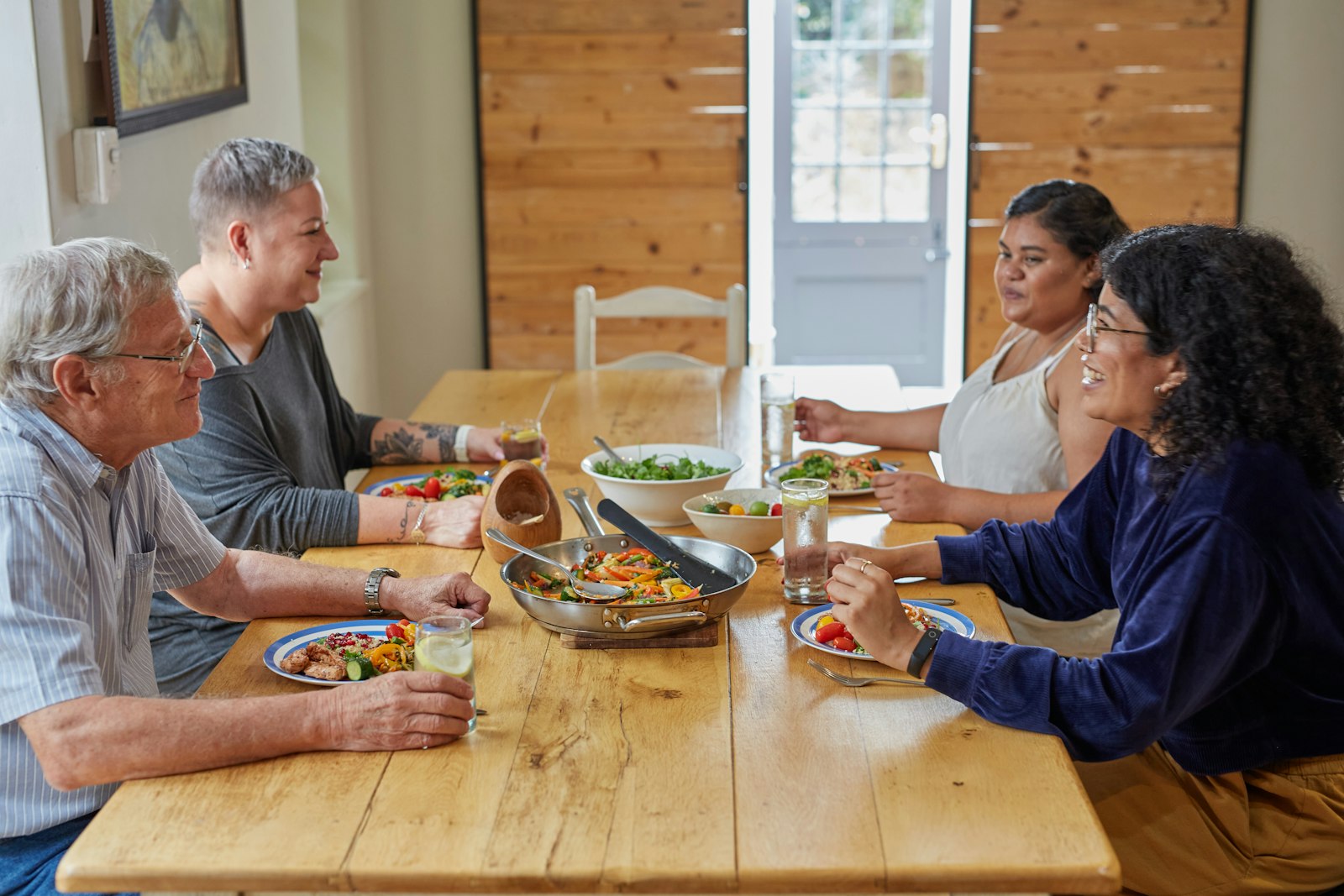 Four adults of different ages seated around a long wooden table sharing a meal of salad, rice and vegetables in a bright room with wooden doors.
