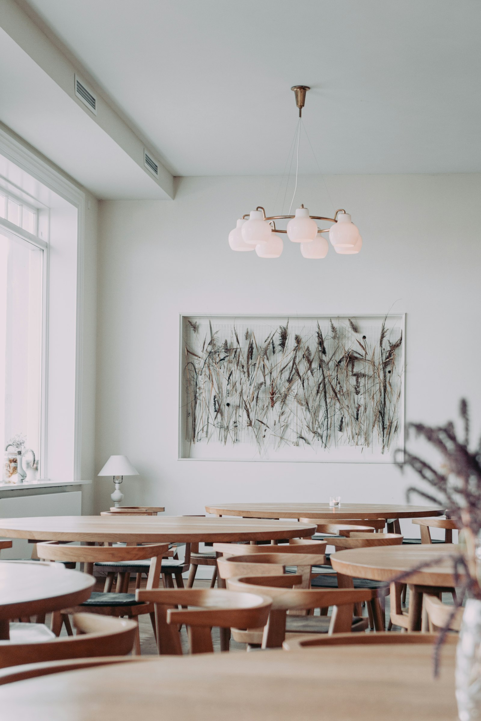A bright dining room with round wooden tables and matching chairs, a framed grass artwork on the wall and a pendant light overhead.