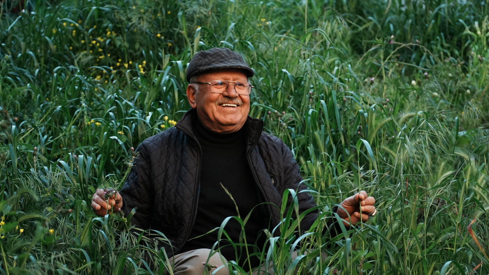 An older man in a flat cap and quilted jacket laughing while seated among tall green meadow grass.