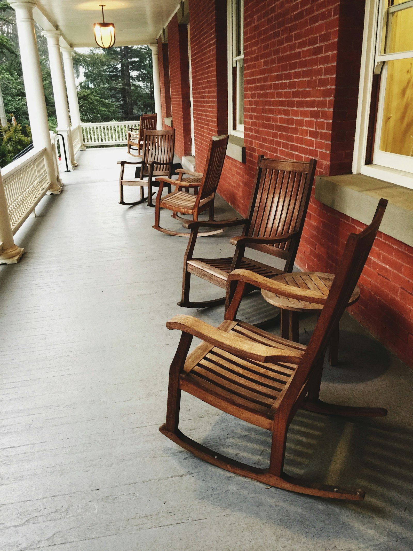 A row of wooden rocking chairs and a small round table along a covered veranda with white columns beside a red brick wall.